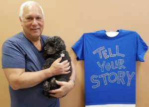 Charles holding Bonnie next to "Tell Your Story" tee-shirt